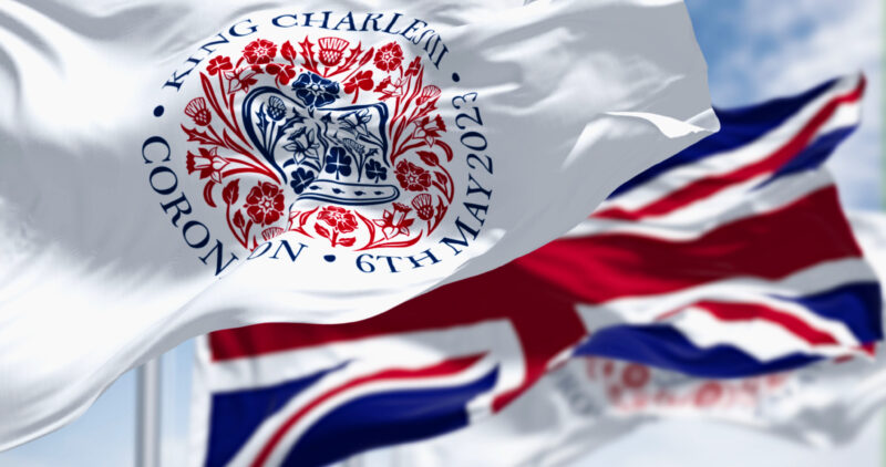 Picture shows_Flags with the emblem of the coronation of King Charles III and of UK waving. The emblem depicts flora of the four nations in the shape of St Edward’s Crown. 3d illustration render. 
(Getty Stock Image_1481418471)
